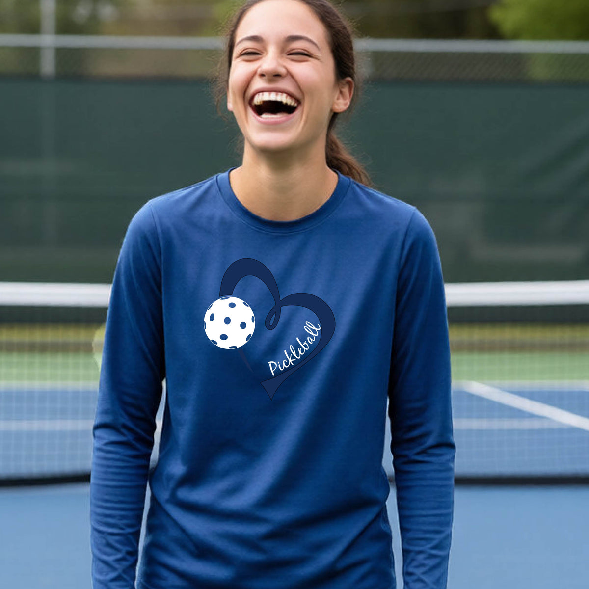 A young person wearing a royal blue long-sleeve athletic t-shirt with a navy blue heart, a white pickleball, and the text 'Pickleball' in script on the front of the shirt. 