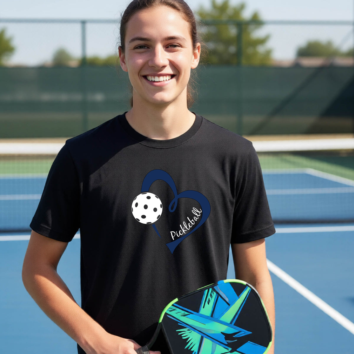 A young person smiling and laughing on a pickleball court, wearing a black t-shirt with a navy blue heart, a white pickleball graphic, and the word 'Pickleball' written on the front.