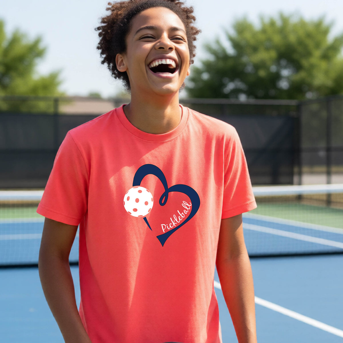 A young person smiling and laughing on a pickleball court, wearing a coral pink t-shirt with a navy blue heart, a white pickleball graphic, and the word 'Pickleball' written on the front.