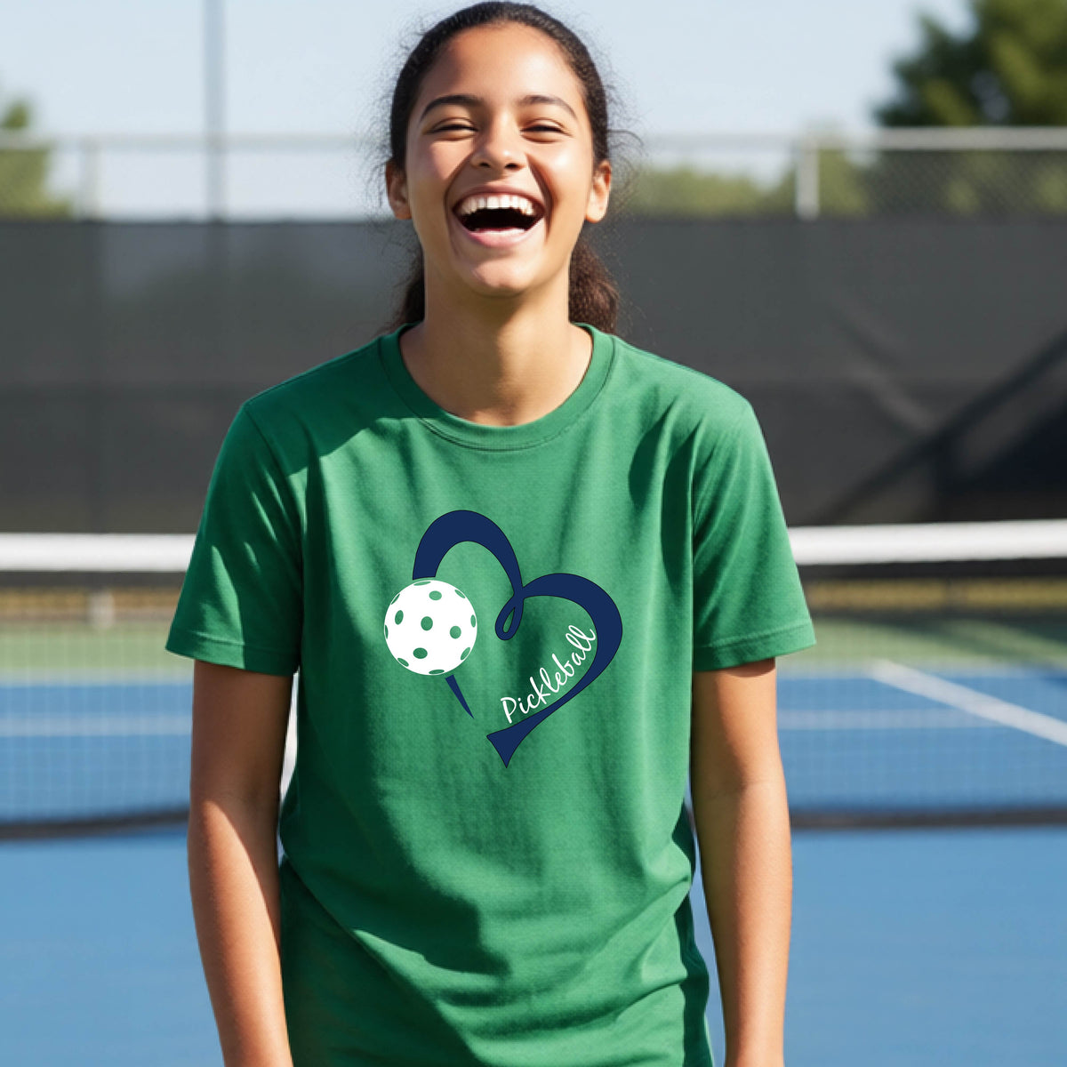 A young person smiling and laughing on a pickleball court, wearing a green t-shirt with a navy blue heart, a white pickleball graphic, and the word 'Pickleball' written on the front.