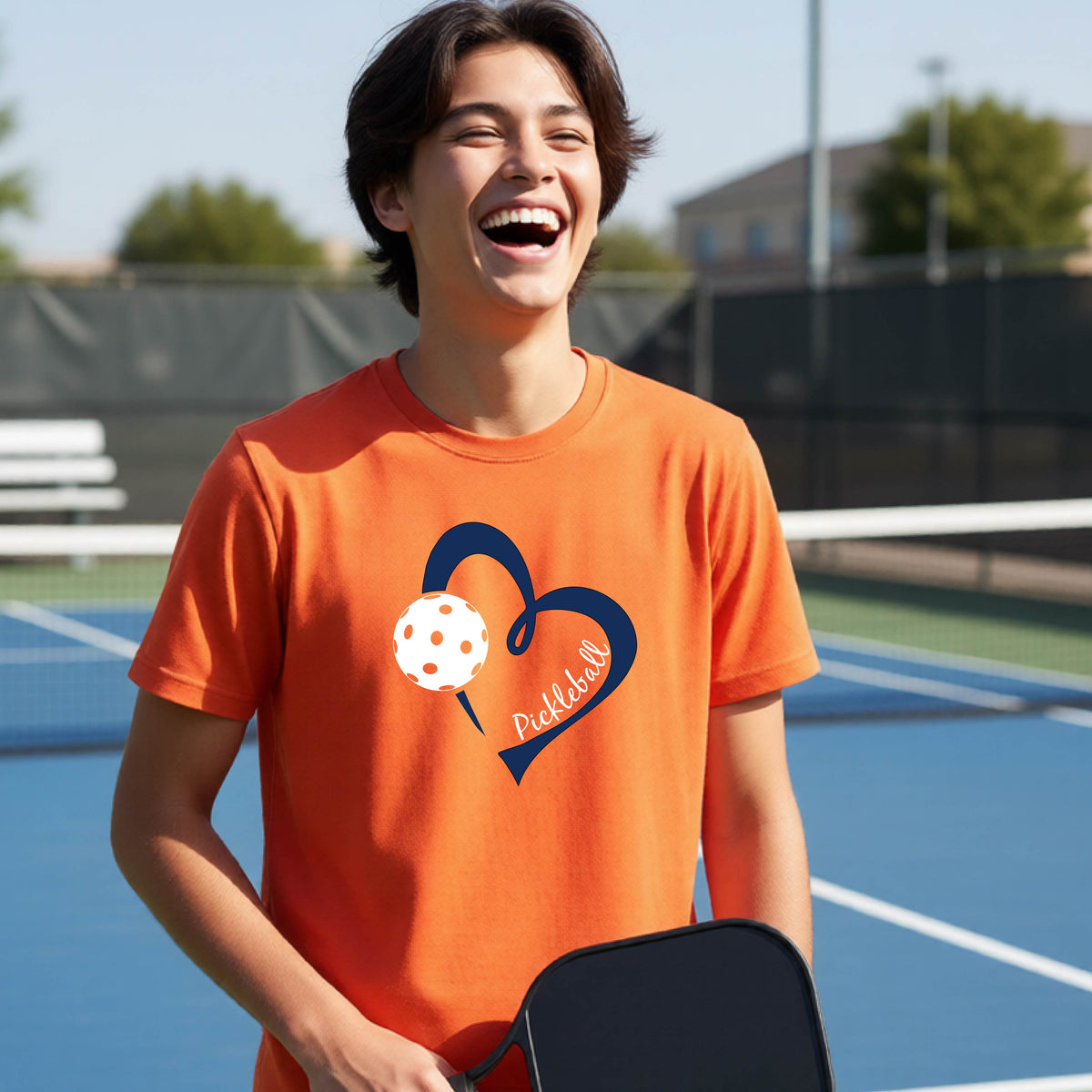 A young person smiling and laughing on a pickleball court, wearing an orange t-shirt with a navy blue heart, a white pickleball graphic, and the word 'Pickleball' written on the front.