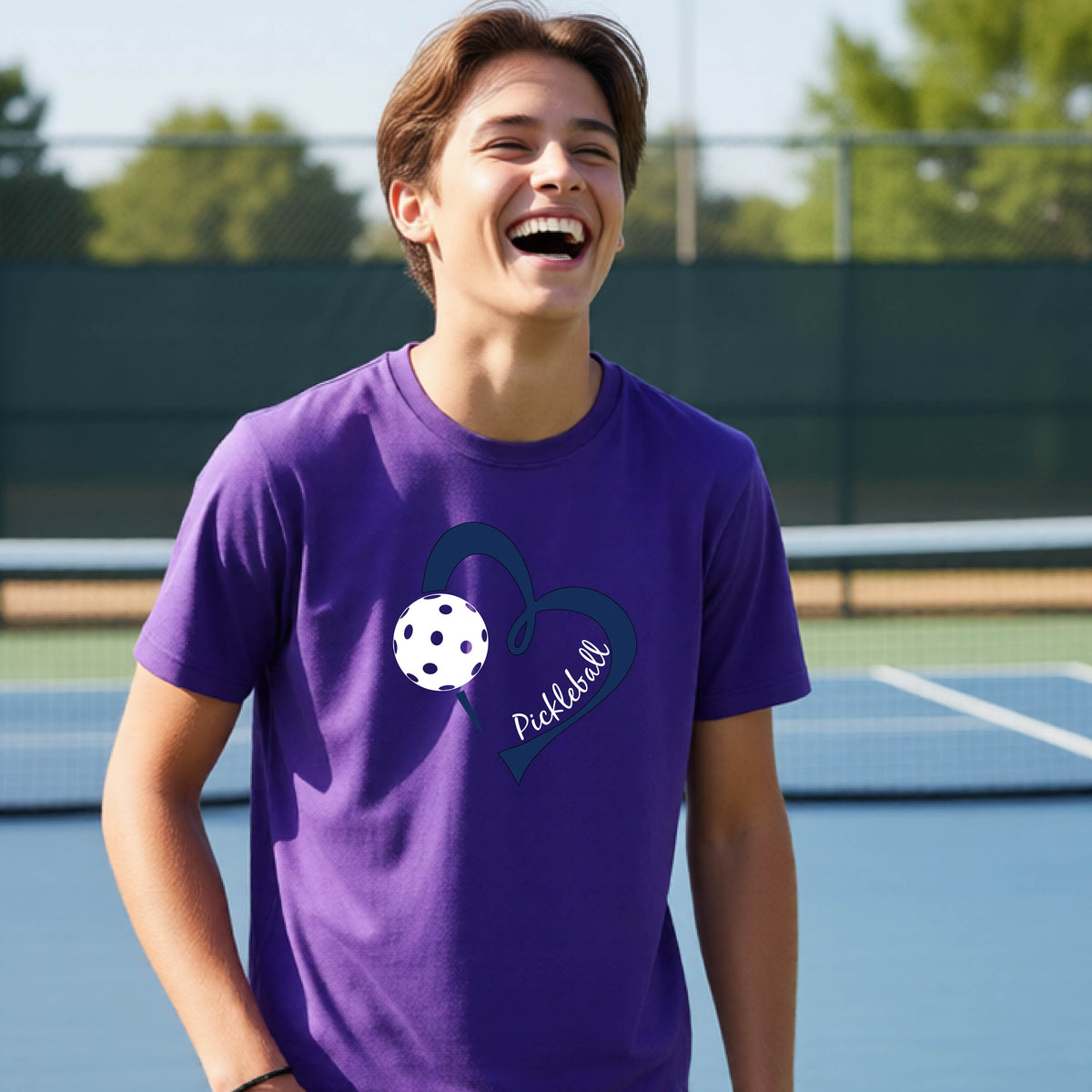 A young person smiling and laughing on a pickleball court, wearing a purple t-shirt with a navy blue heart, a white pickleball graphic, and the word 'Pickleball' written on the front.