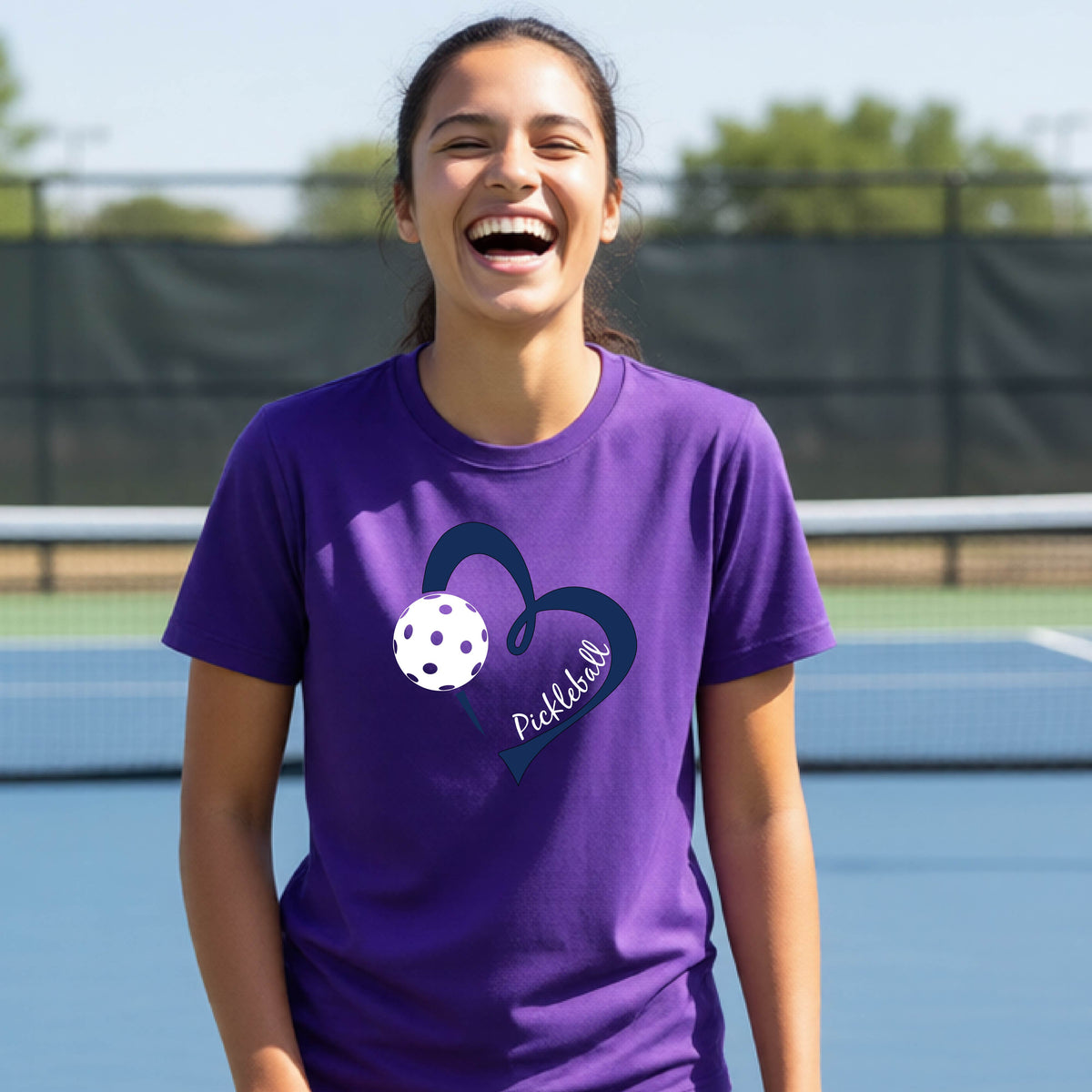 A young person smiling and laughing on a pickleball court, wearing a purple t-shirt with a navy blue heart, a white pickleball graphic, and the word 'Pickleball' written on the front.