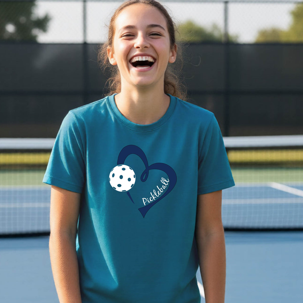 A young person smiling and laughing on a pickleball court, wearing a teal t-shirt with a navy blue heart, a white pickleball graphic, and the word 'Pickleball' written on the front.
