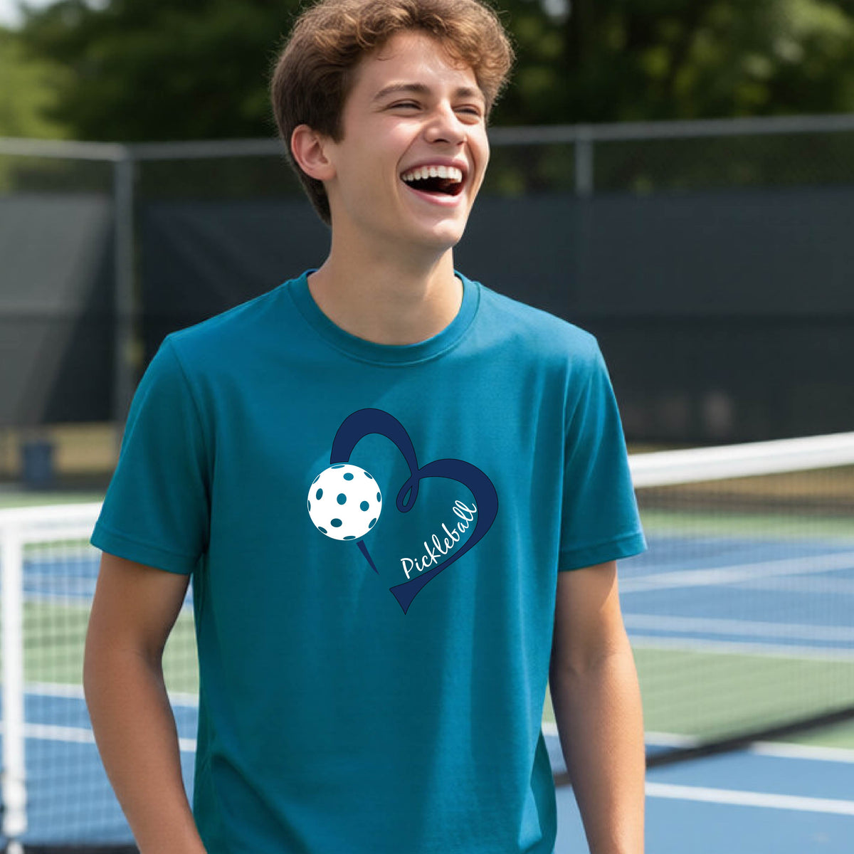 A young person smiling and laughing on a pickleball court, wearing a teal t-shirt with a navy blue heart, a white pickleball graphic, and the word 'Pickleball' written on the front.