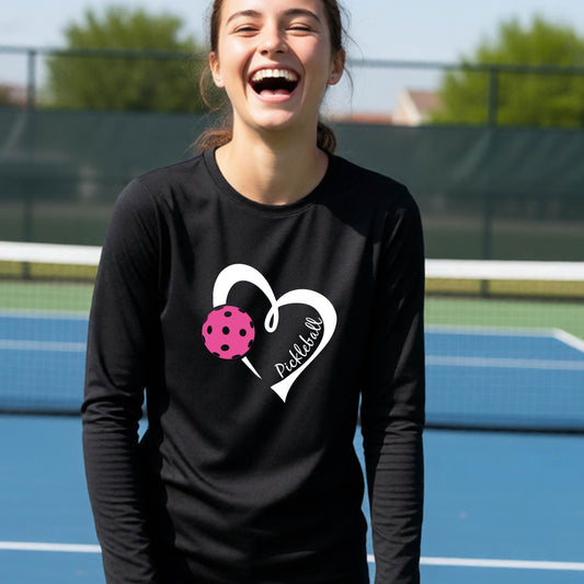 A young person wearing a black long-sleeve t-shirt with a navy blue heart, pink pickleball icon and the text "Pickleball' written in script on the front. 