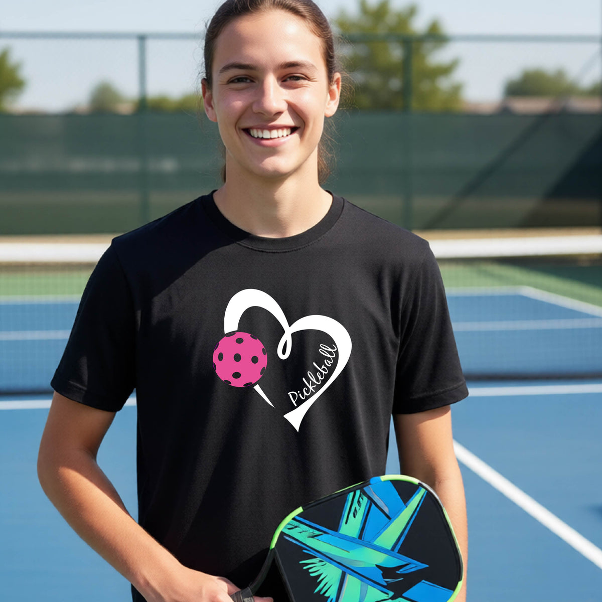 A teenager laughing on an outdoor pickleball court wearing a black t-shirt with a white heart graphic, a pink pickleball, and the text 'Pickleball' inside the heart. 