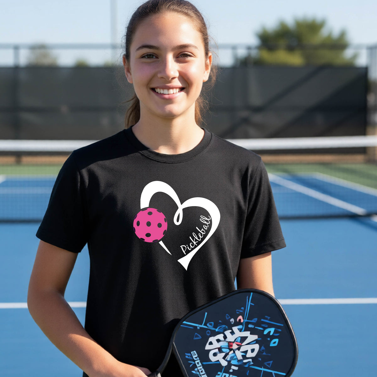 A teenager laughing on an outdoor pickleball court wearing a black t-shirt with a white heart graphic, a pink pickleball, and the text 'Pickleball' inside the heart. 