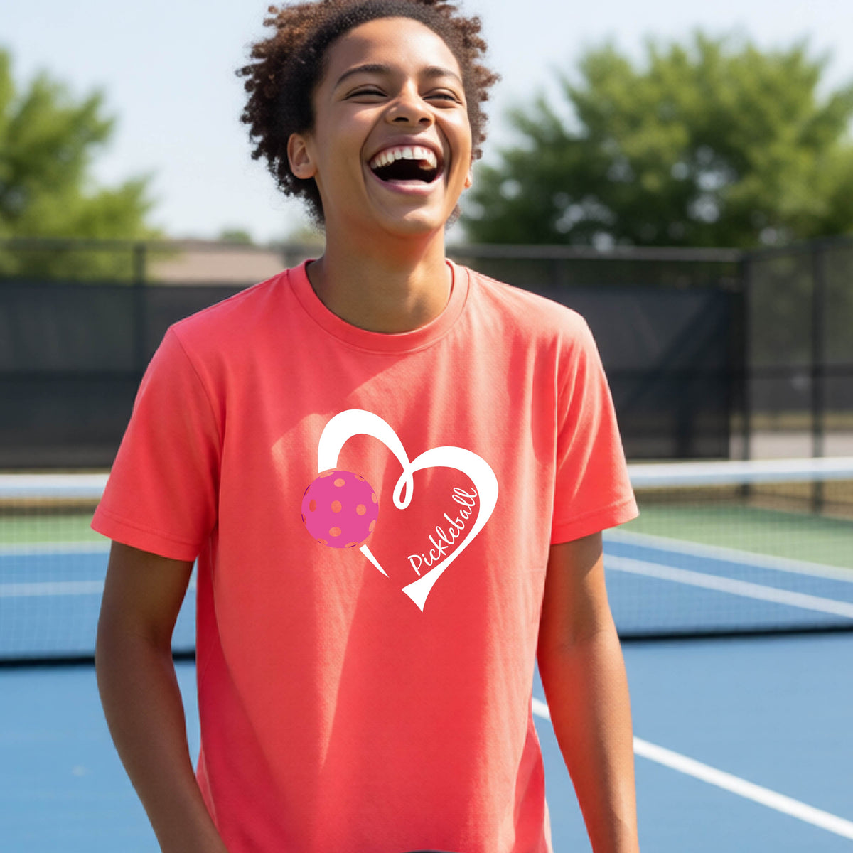 A teenager laughing on an outdoor pickleball court wearing a coral pink t-shirt with a white heart graphic, a pink pickleball, and the text 'Pickleball' inside the heart. 