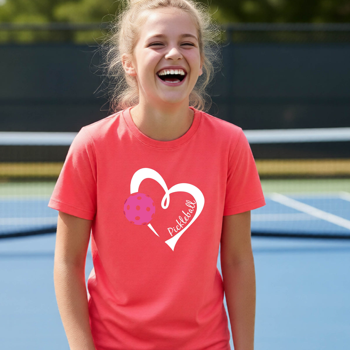 A teenager laughing on an outdoor pickleball court wearing a coral pink t-shirt with a white heart graphic, a pink pickleball, and the text 'Pickleball' inside the heart. 