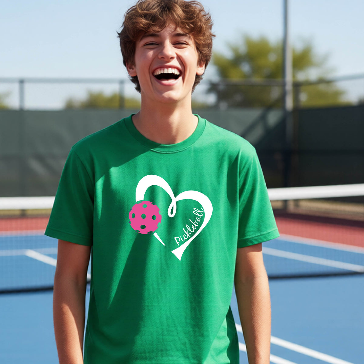 A teenager laughing on an outdoor pickleball court wearing a green t-shirt with a white heart graphic, a pink pickleball, and the text 'Pickleball' inside the heart. 