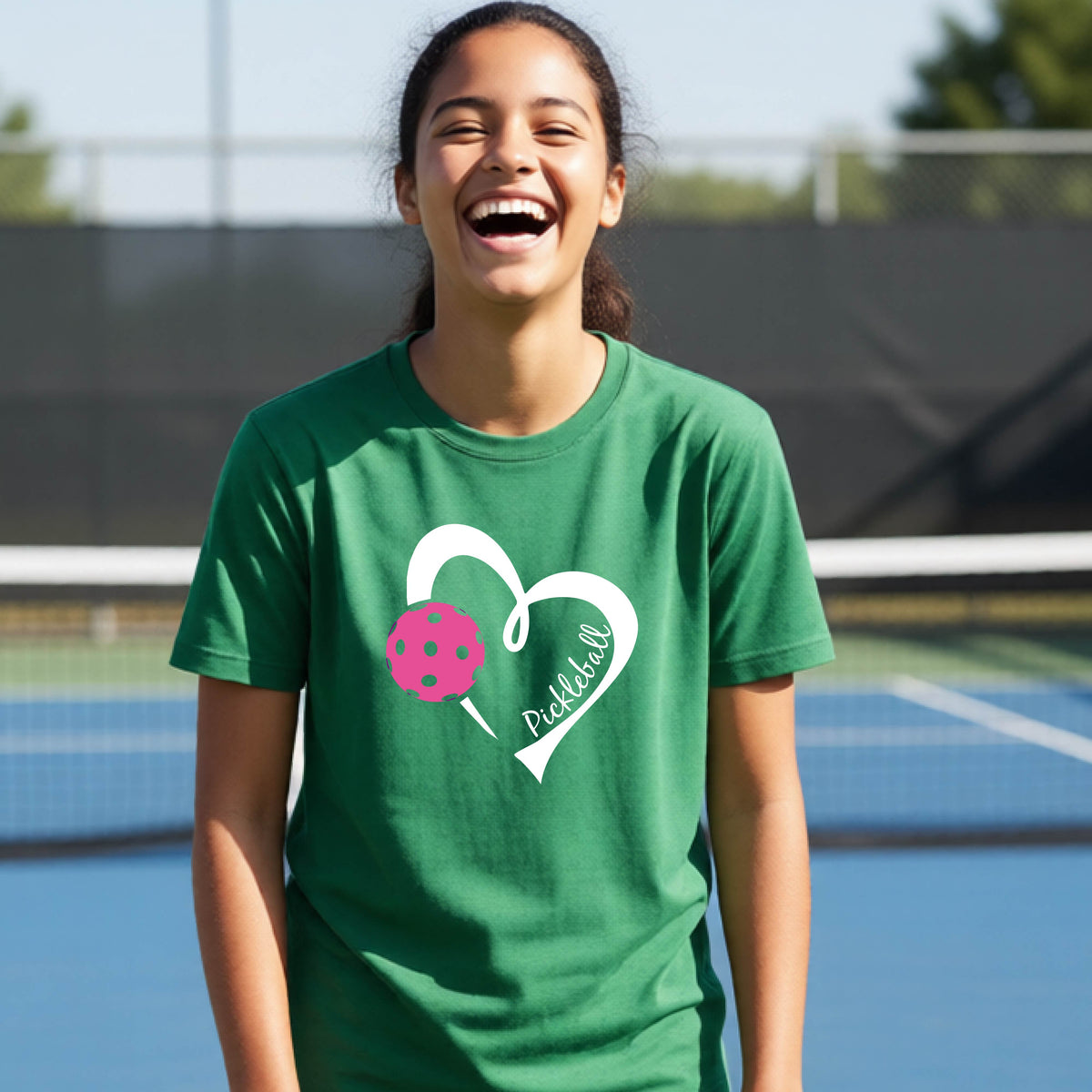 A teenager laughing on an outdoor pickleball court wearing a green t-shirt with a white heart graphic, a pink pickleball, and the text 'Pickleball' inside the heart. 