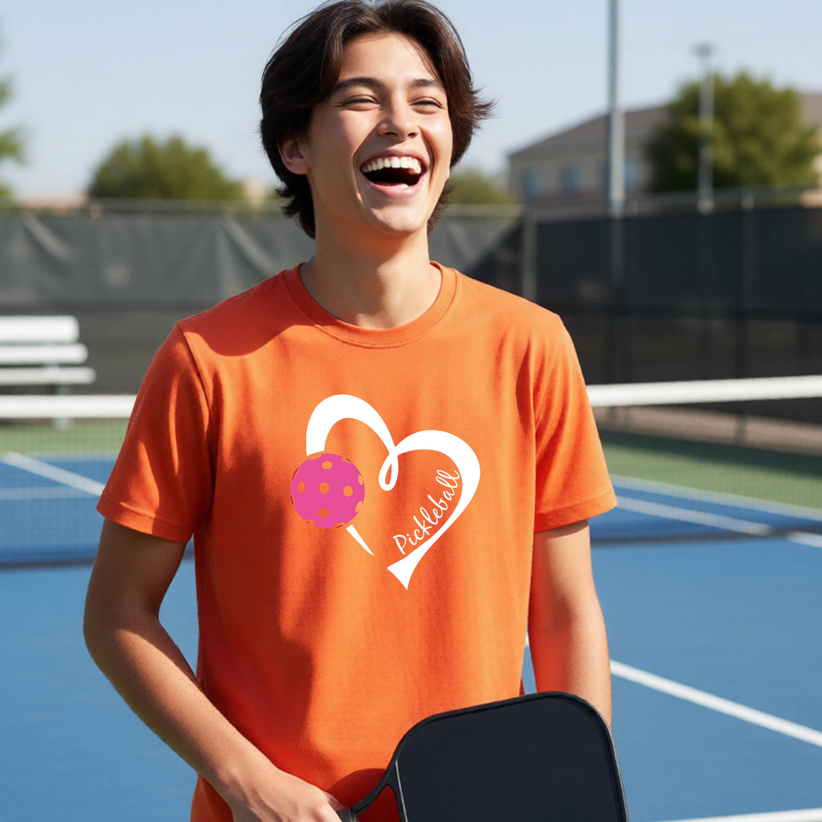 A teenager laughing on an outdoor pickleball court wearing an orange t-shirt with a white heart graphic, a pink pickleball, and the text 'Pickleball' inside the heart. 
