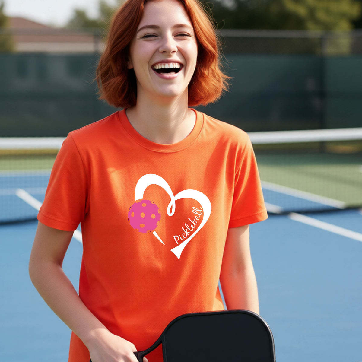 A teenager laughing on an outdoor pickleball court wearing an orange t-shirt with a white heart graphic, a pink pickleball, and the text 'Pickleball' inside the heart. 