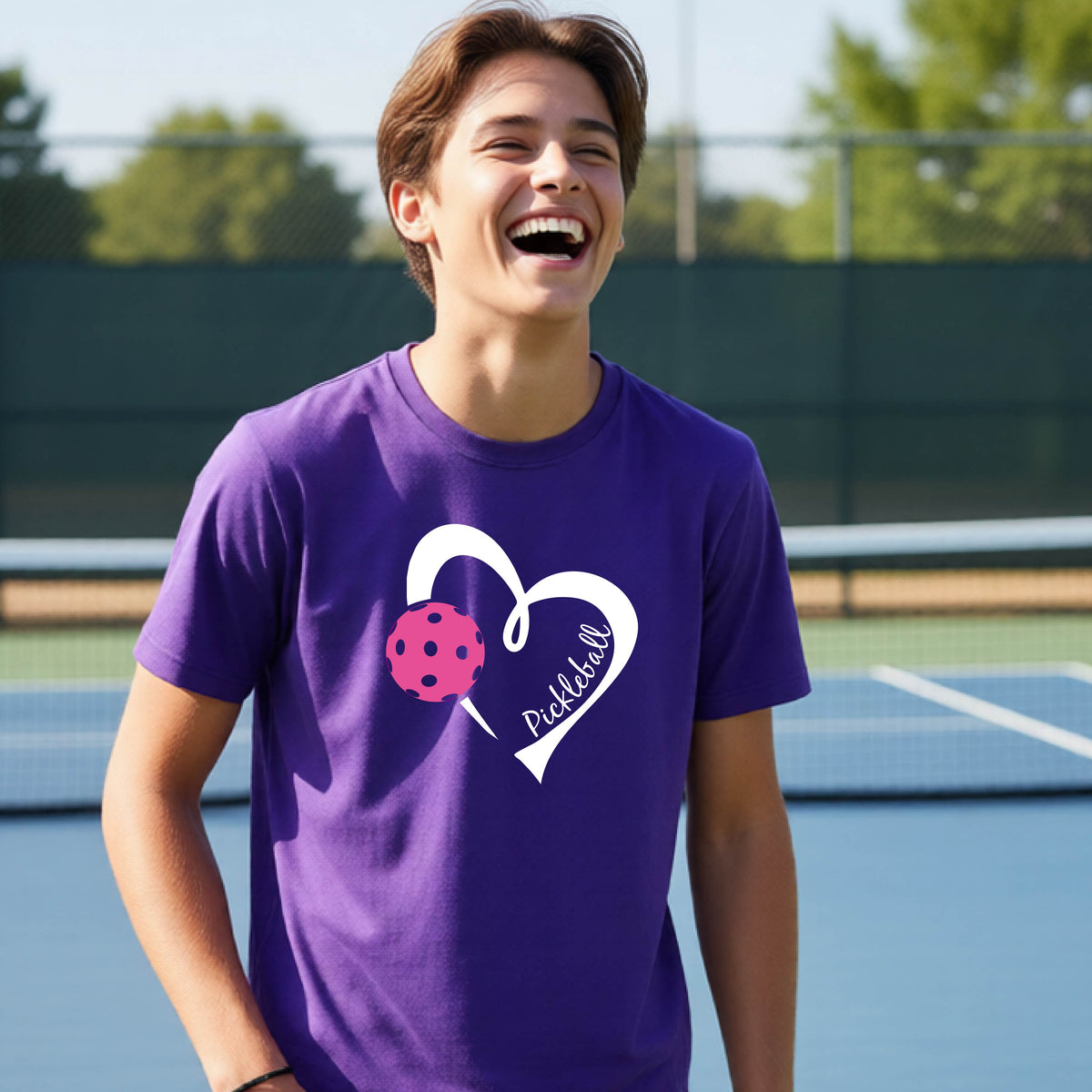 A teenager laughing on an outdoor pickleball court wearing a purple t-shirt with a white heart graphic, a pink pickleball, and the text 'Pickleball' inside the heart. 