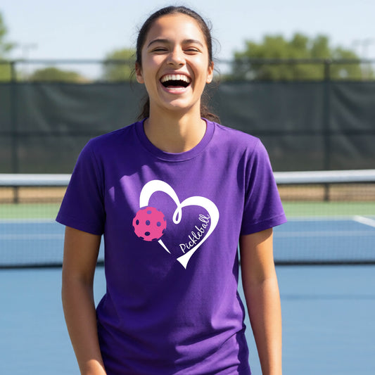 A teenager laughing on an outdoor pickleball court wearing a purple t-shirt with a white heart graphic, a pink pickleball, and the text 'Pickleball' inside the heart. 