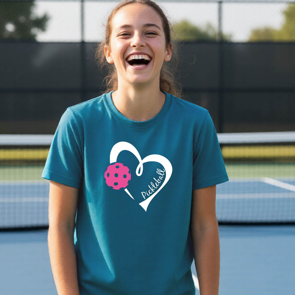 A teenager laughing on an outdoor pickleball court wearing a teal t-shirt with a white heart graphic, a pink pickleball, and the text 'Pickleball' inside the heart. 