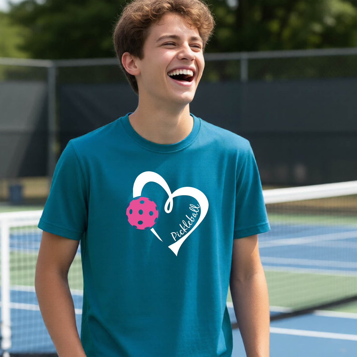 A teenager laughing on an outdoor pickleball court wearing a teal t-shirt with a white heart graphic, a pink pickleball, and the text 'Pickleball' inside the heart. 