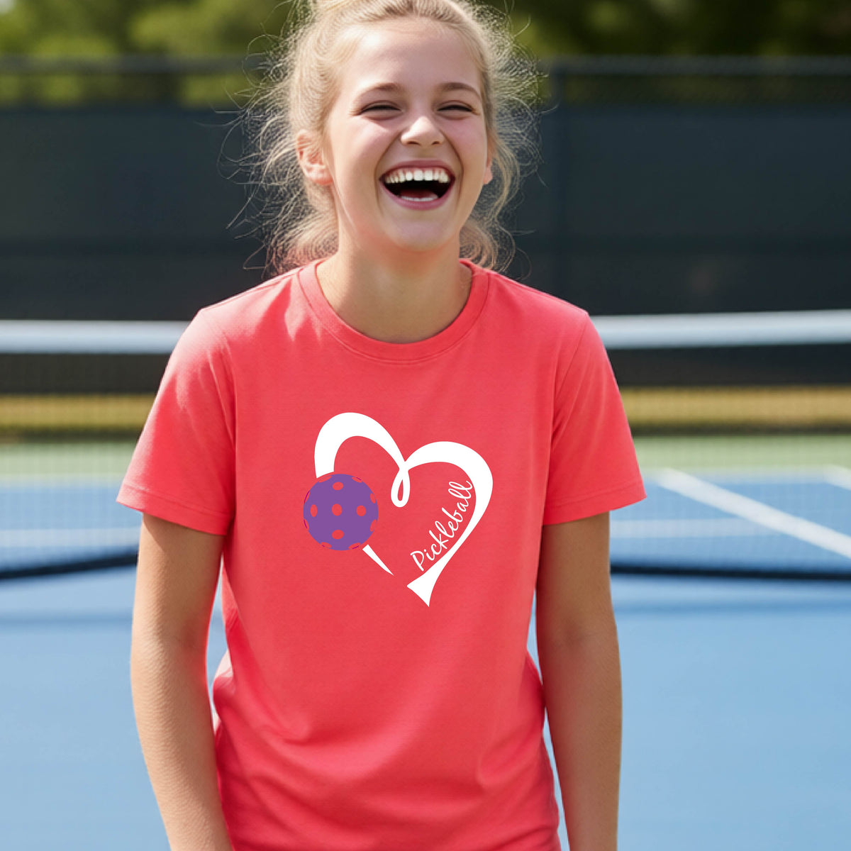 A young person laughing on an outdoor pickleball court wearing a coral pink t-shirt with a white heart graphic, a purple pickleball, and the text 'Pickleball' on the front. 