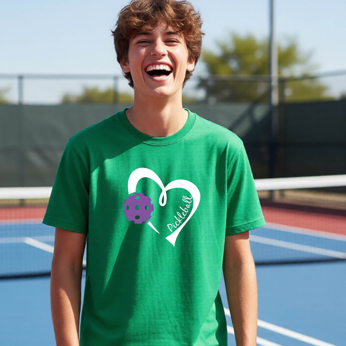 A young person laughing on an outdoor pickleball court wearing a green t-shirt with a white heart graphic, a purple pickleball, and the text 'Pickleball' on the front. 