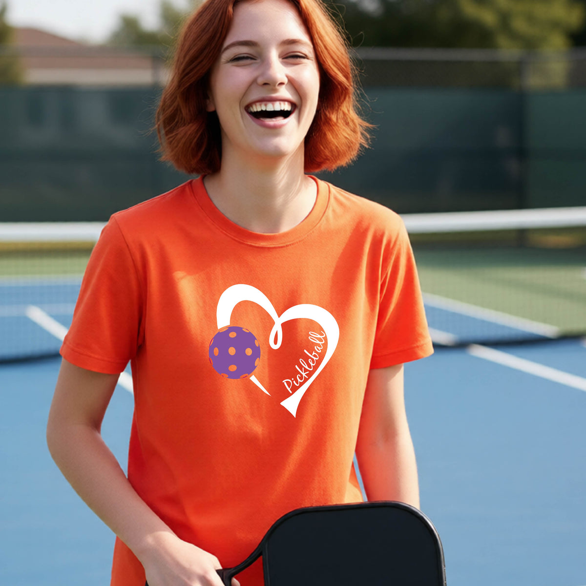 A young person laughing on an outdoor pickleball court wearing a orange t-shirt with a white heart graphic, a purple pickleball, and the text 'Pickleball' on the front. 