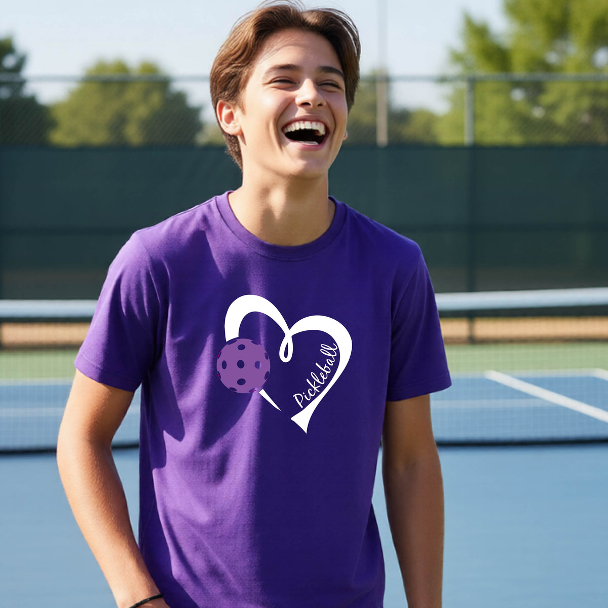 A young person laughing on an outdoor pickleball court wearing a purple t-shirt with a white heart graphic, a purple pickleball, and the text 'Pickleball' on the front. 