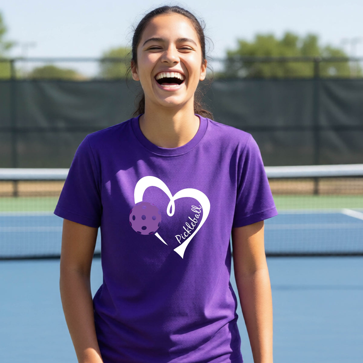 A young person laughing on an outdoor pickleball court wearing a purple t-shirt with a white heart graphic, a purple pickleball, and the text 'Pickleball' on the front. 