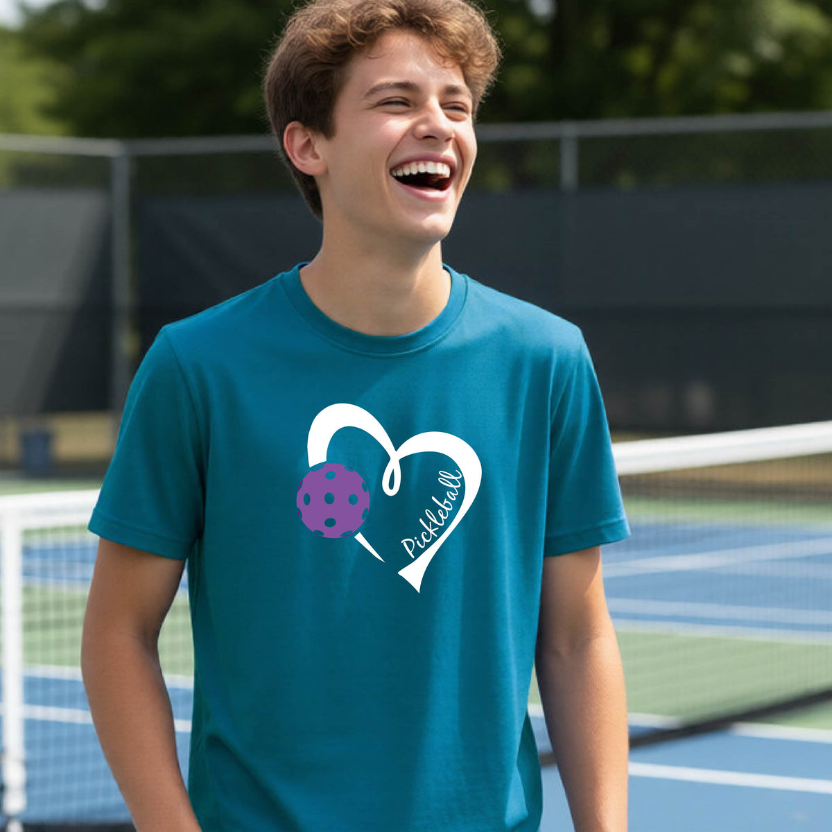 A young person laughing on an outdoor pickleball court wearing a teal t-shirt with a white heart graphic, a purple pickleball, and the text 'Pickleball' on the front. 