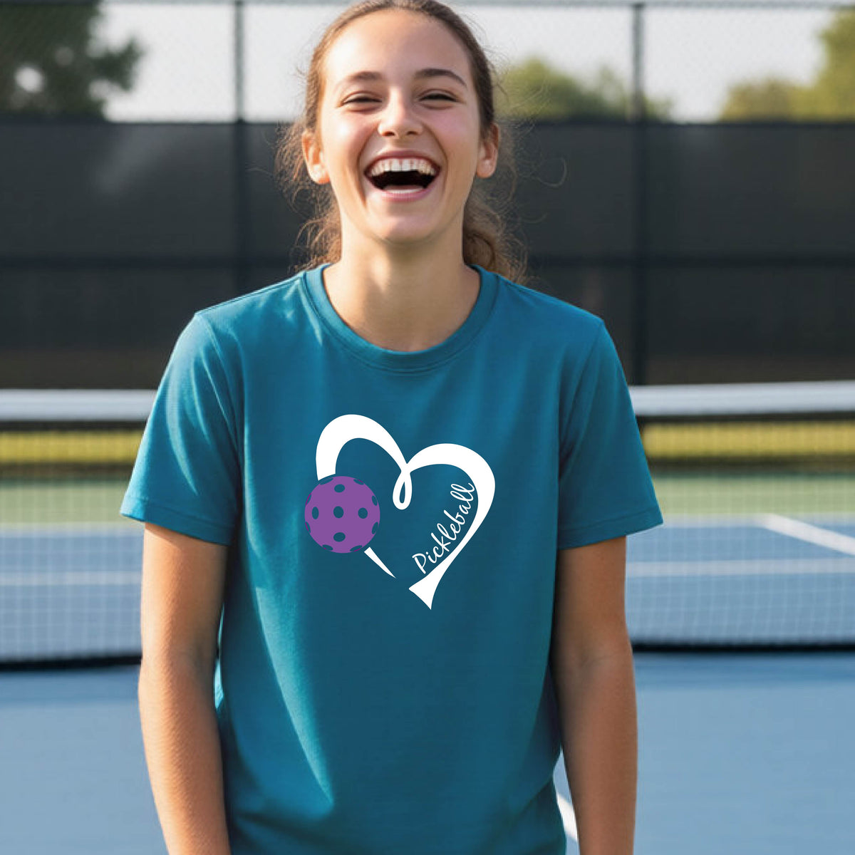 A young person laughing on an outdoor pickleball court wearing a teal t-shirt with a white heart graphic, a purple pickleball, and the text 'Pickleball' on the front. 