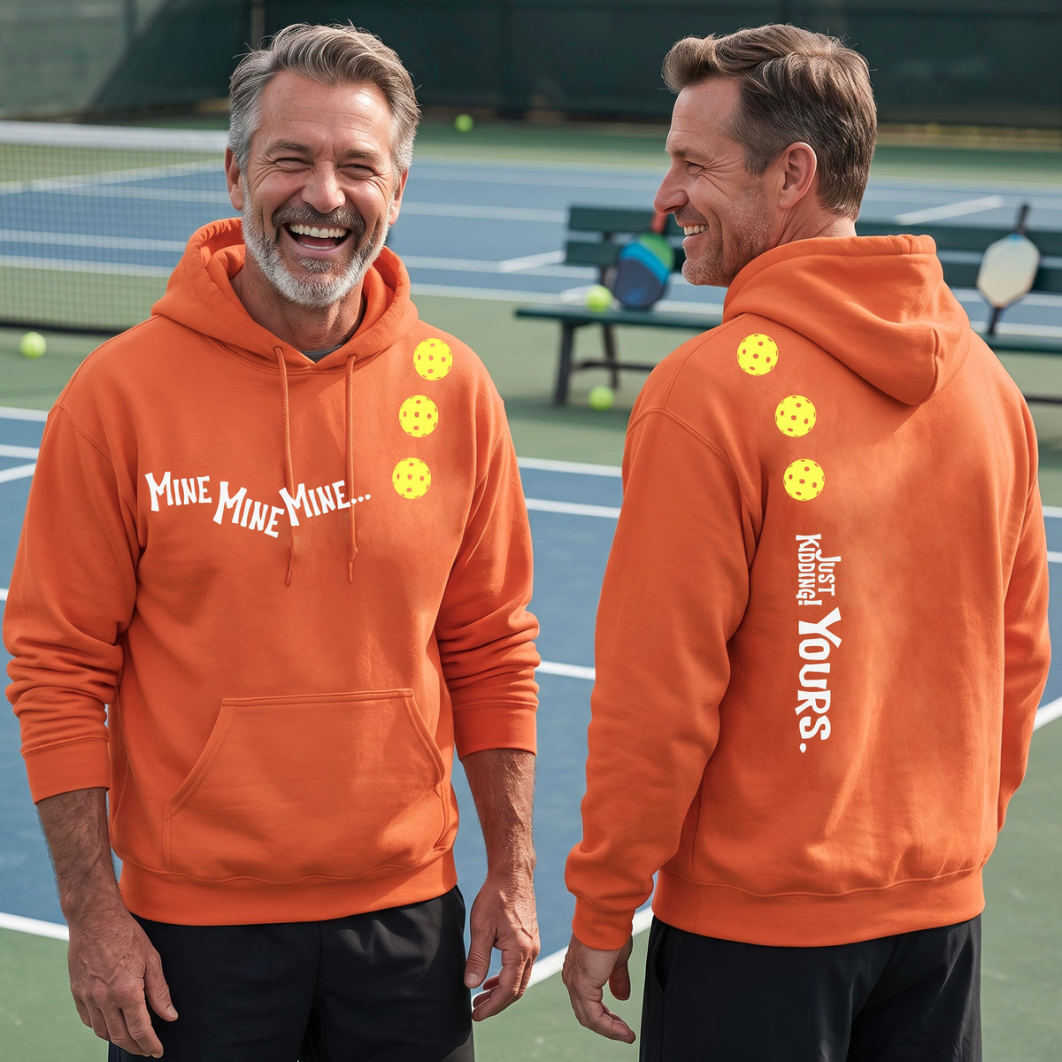 Two men on a pickleball court wearing matching bright orange unisex hoodie pullovers. One shows the front with the text 'MINE MINE MINE' along with three pickleball graphics. and the back text 'JUST KIDDING YOURS, along with three pickleball graphics.