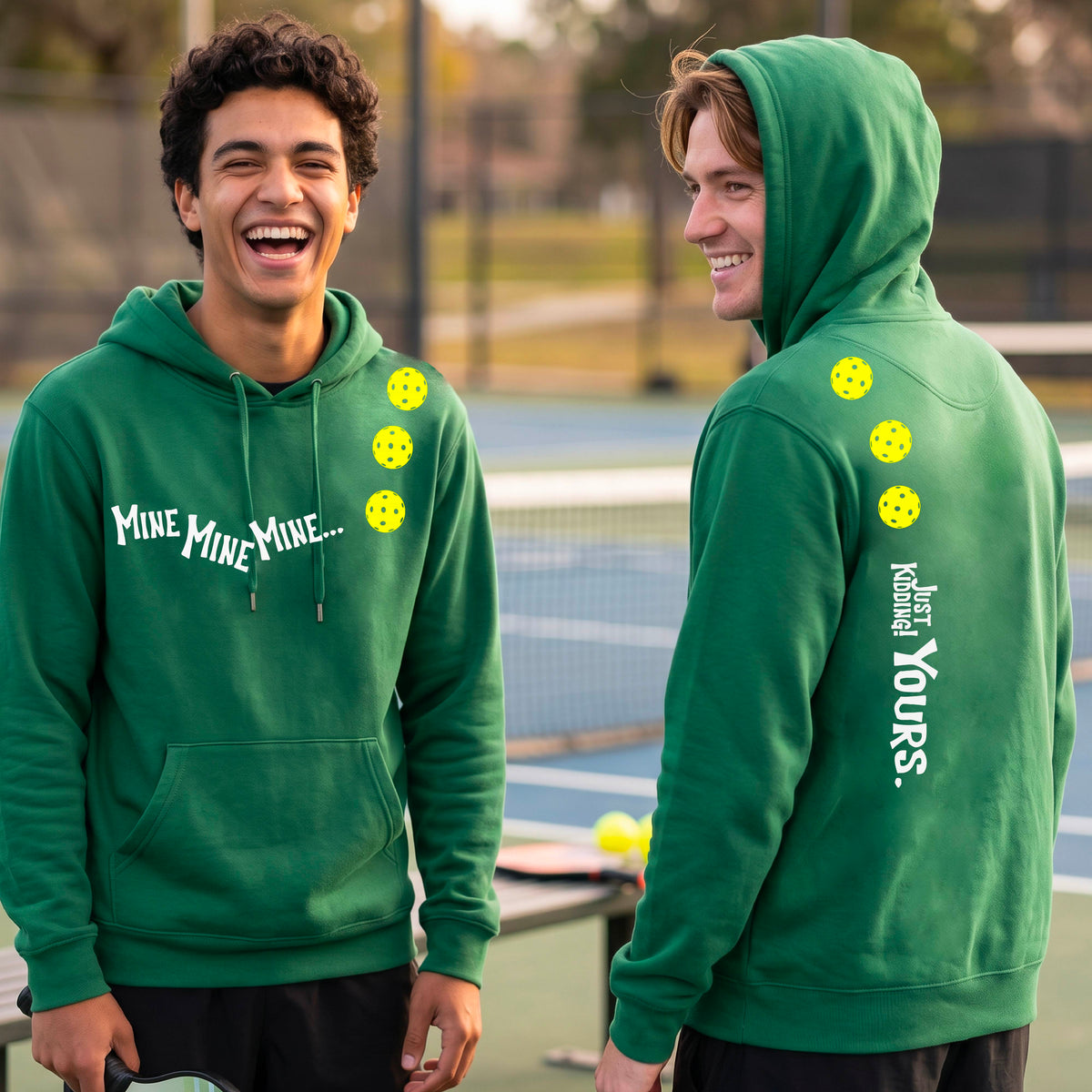 Two men on a pickleball court wearing matching green unisex hoodie pullovers. One shows the front with the text 'MINE MINE MINE' along with three pickleball graphics. and the back text 'JUST KIDDING YOURS, along with three pickleball graphics.