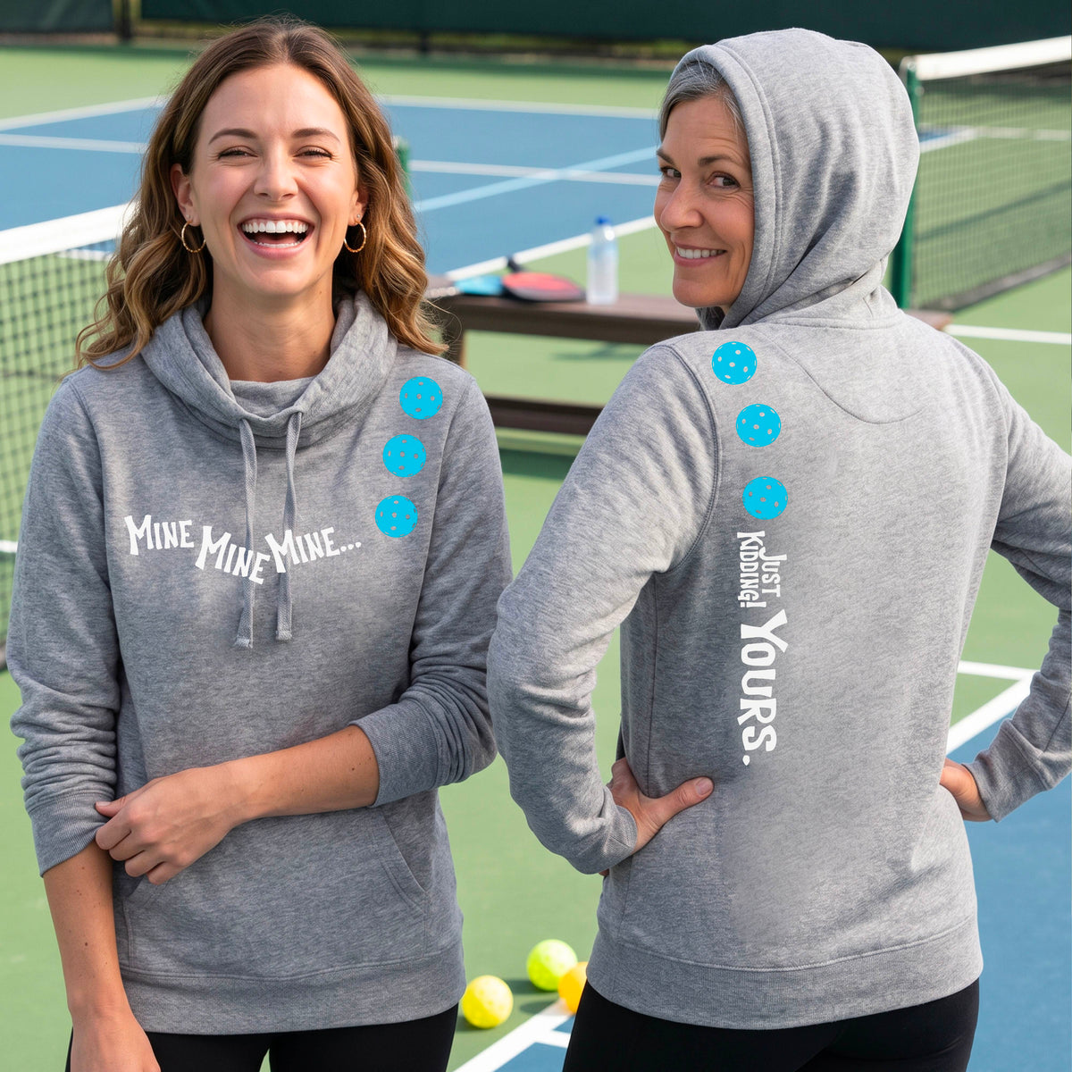 Two women on a pickleball court wearing matching cowl-neck gray hoodies One shows the front with the text 'MINE MINE MINE' along with three pickleball graphics. and the back text 'JUST KIDDING YOURS, along with three pickleball graphics.
