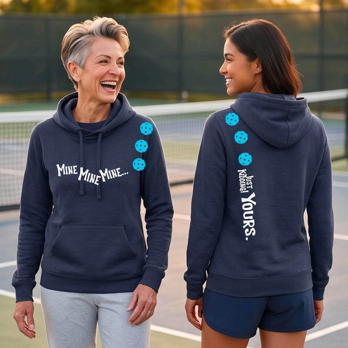 Two women on a pickleball court wearing matching cowl-neck navy-blue hoodies One shows the front with the text 'MINE MINE MINE' along with three pickleball graphics. and the back text 'JUST KIDDING YOURS, along with three pickleball graphics.