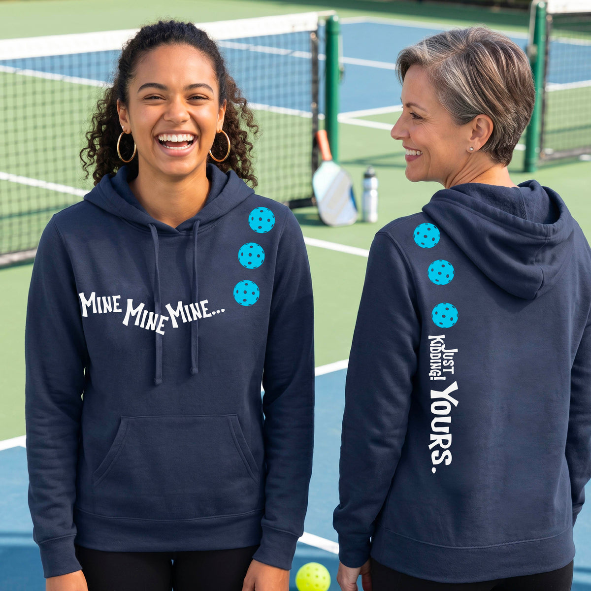 Two women on a pickleball court wearing matching navy blue fitted hoodie pullovers. One shows the front with the text 'MINE MINE MINE' along with three pickleball graphics. and the back text 'JUST KIDDING YOURS, along with three pickleball graphics.