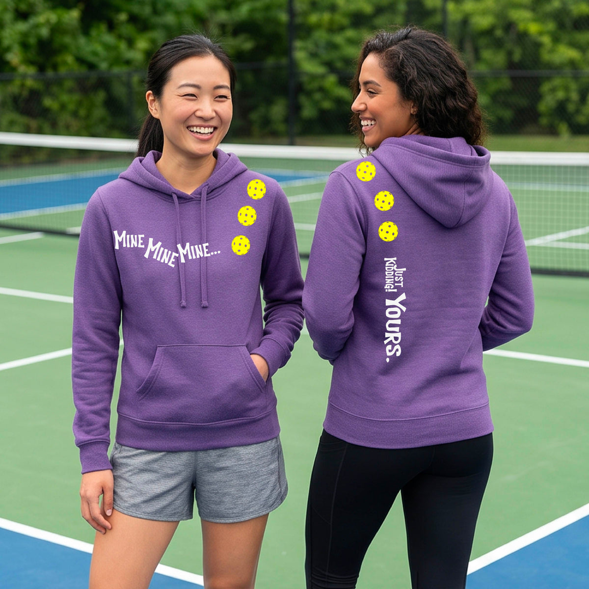 Two women on a pickleball court wearing matching purple fitted hoodie pullovers. One shows the front with the text 'MINE MINE MINE' along with three pickleball graphics. and the back text 'JUST KIDDING YOURS, along with three pickleball graphics.