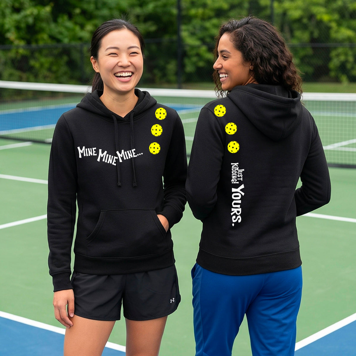 Two women on a pickleball court wearing matching black fitted hoodie pullovers. One shows the front with the text 'MINE MINE MINE' along with three pickleball graphics. and the back text 'JUST KIDDING YOURS, along with three pickleball graphics.