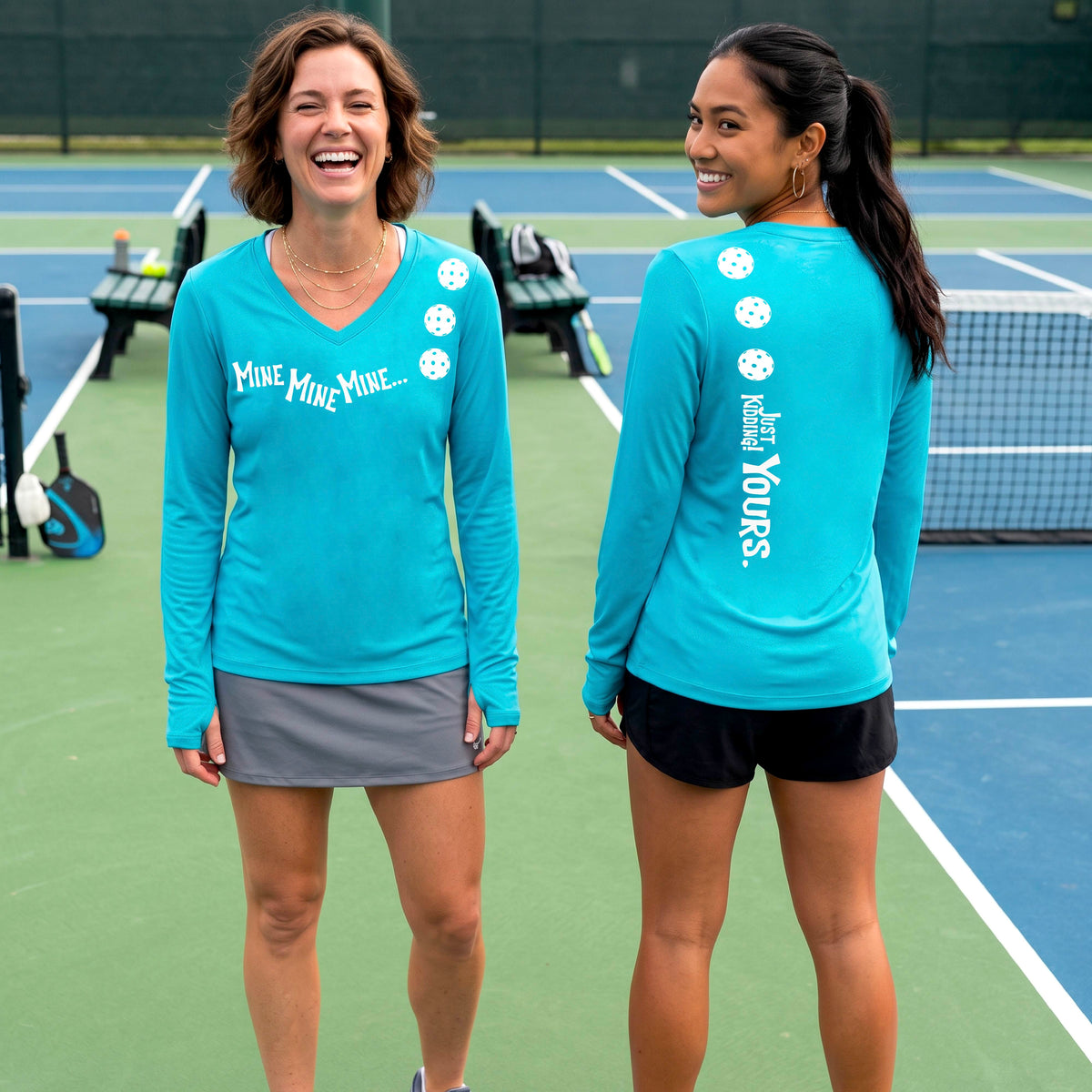 Two women on a pickleball court wearing matching bright blue long-sleeve athletic t-shirts. One shows the front with the text 'MINE MINE MINE' along with three pickleball graphics. and the back text 'JUST KIDDING YOURS, along with three pickleball graphics.