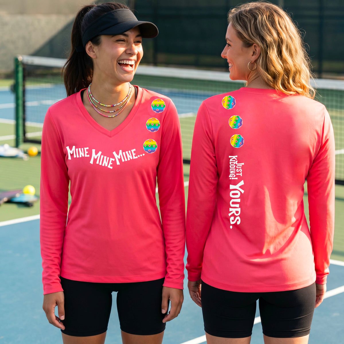 Two women on a pickleball court wearing matching coral pink long-sleeve athletic t-shirts. One shows the front with the text 'MINE MINE MINE' along with three pickleball graphics. and the back text 'JUST KIDDING YOURS, along with three pickleball graphics.