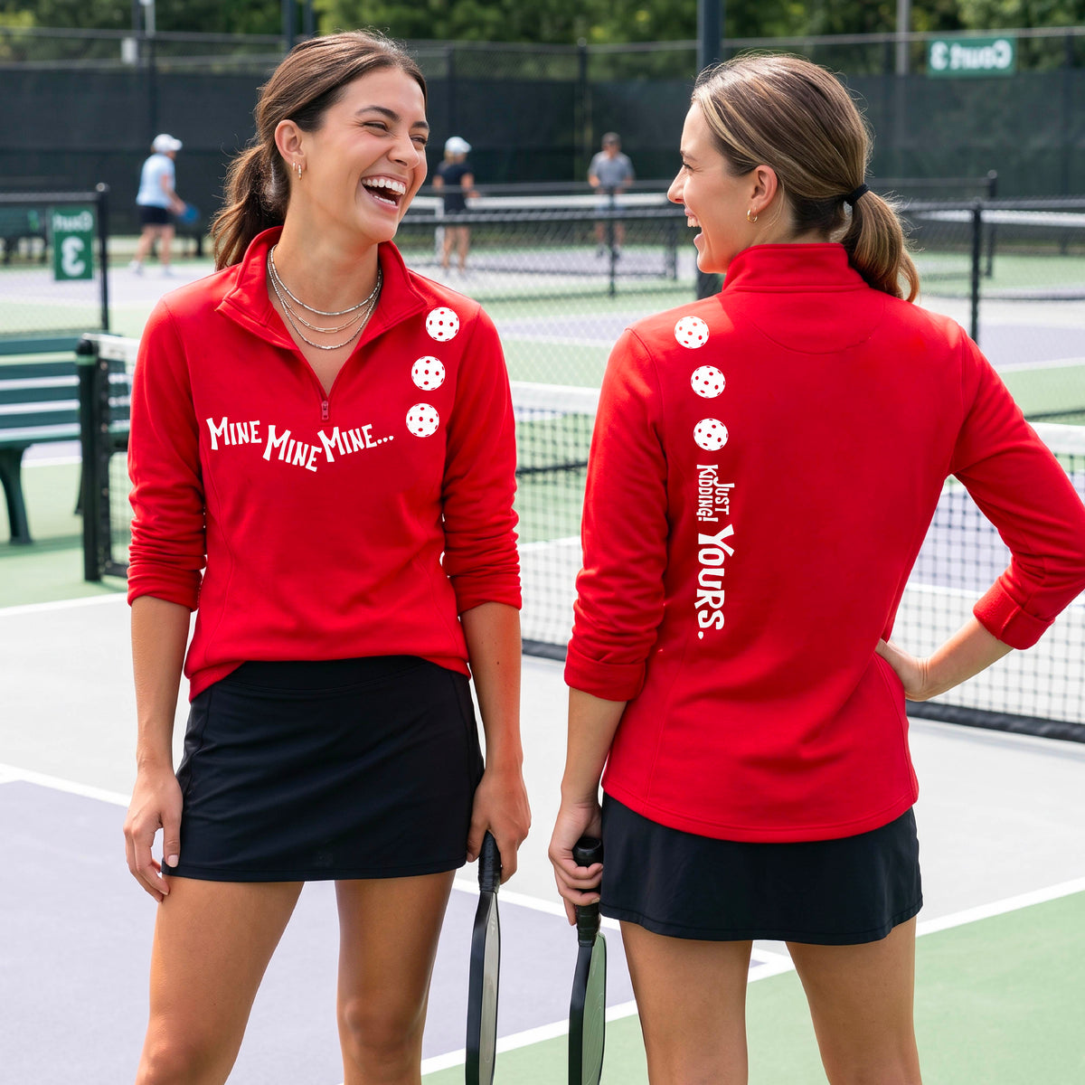 Two women on a pickleball court wearing matching red quarter-zip pullover shirts. One shows the front with the text 'MINE MINE MINE' along with three pickleball graphics. and the back text 'JUST KIDDING YOURS, along with three pickleball graphics.