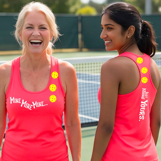 Two women on a pickleball court wearing matching bright coral pink athletic tank tops  One shows the front with the text 'MINE MINE MINE' along with three pickleball graphics. and the back text 'JUST KIDDING YOURS, along with three pickleball graphics.