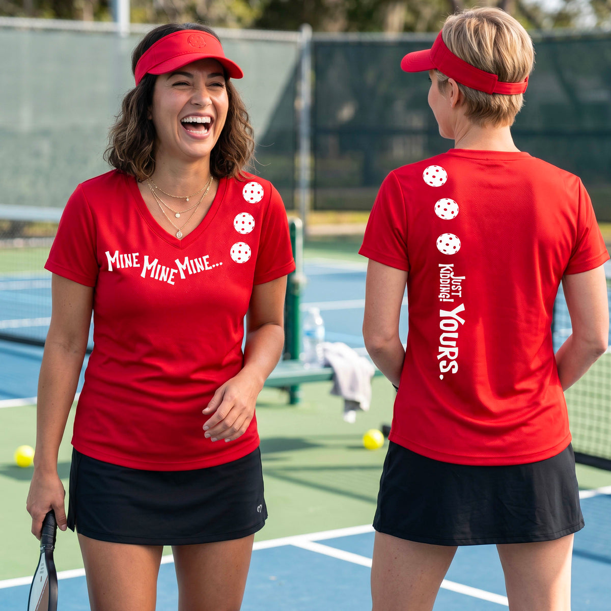 Two women on a pickleball court wearing matching red short-sleeve v-neck t-shirts.  One shows the front with the text 'MINE MINE MINE' along with three pickleball graphics. and the back text 'JUST KIDDING YOURS, along with three pickleball graphics.