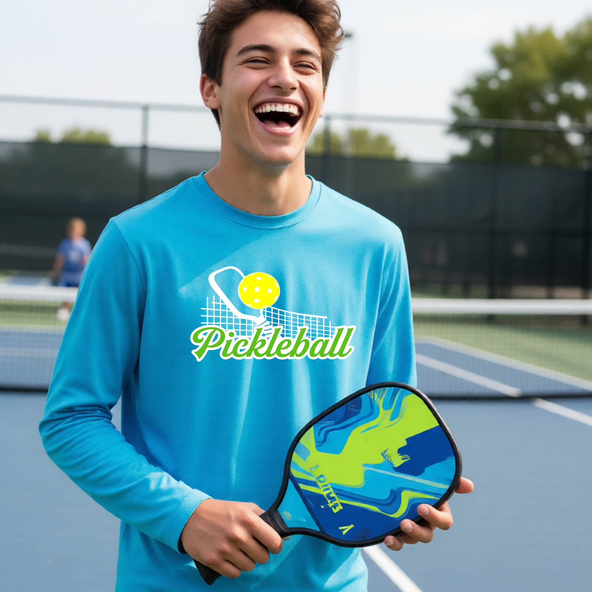 A young person wearing a bright blue long sleeve t-shirt with a graphic that has white and green text "PICKLEBALL'  with a white pickleball paddle and net and a yellow pickleball above the text.