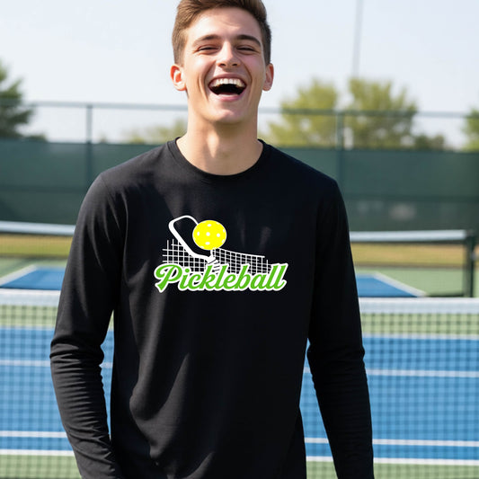 A young person wearing a black long sleeve t-shirt with a graphic that has white and green text "PICKLEBALL'  with a white pickleball paddle and net and a yellow pickleball above the text.