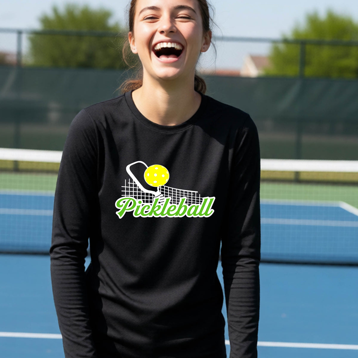 A young person wearing a black long sleeve t-shirt with a graphic that has white and green text "PICKLEBALL'  with a white pickleball paddle and net and a yellow pickleball above the text.