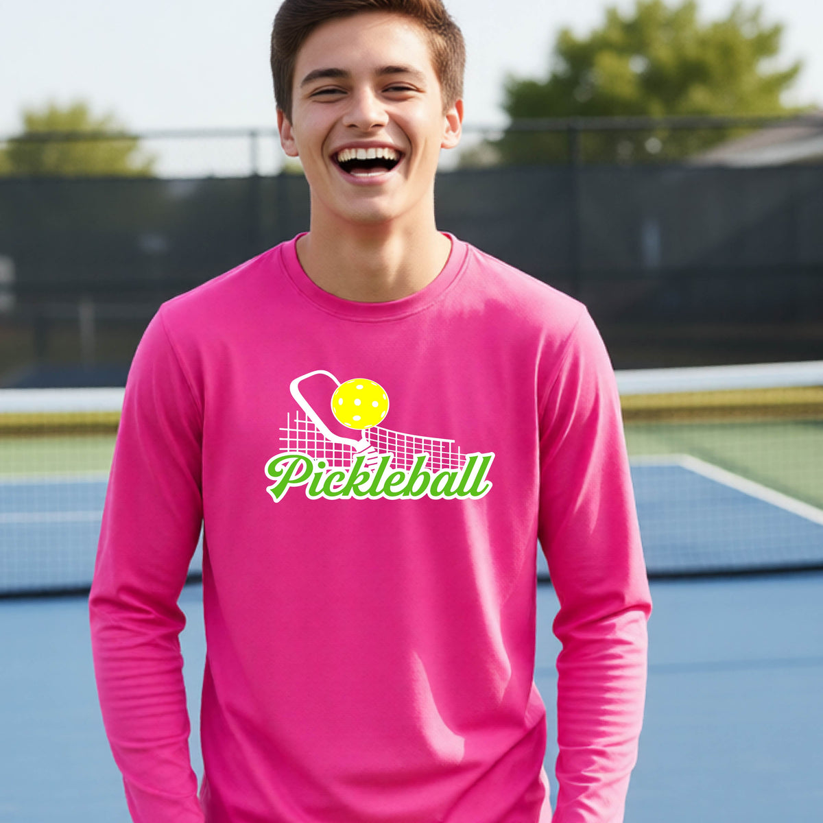 A young person wearing a pink long sleeve t-shirt with a graphic that has white and green text "PICKLEBALL'  with a white pickleball paddle and net and a yellow pickleball above the text.