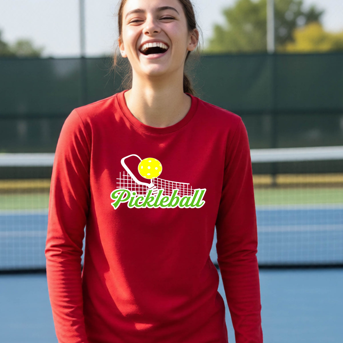 A young person wearing a red long sleeve t-shirt with a graphic that has white and green text "PICKLEBALL'  with a white pickleball paddle and net and a yellow pickleball above the text.