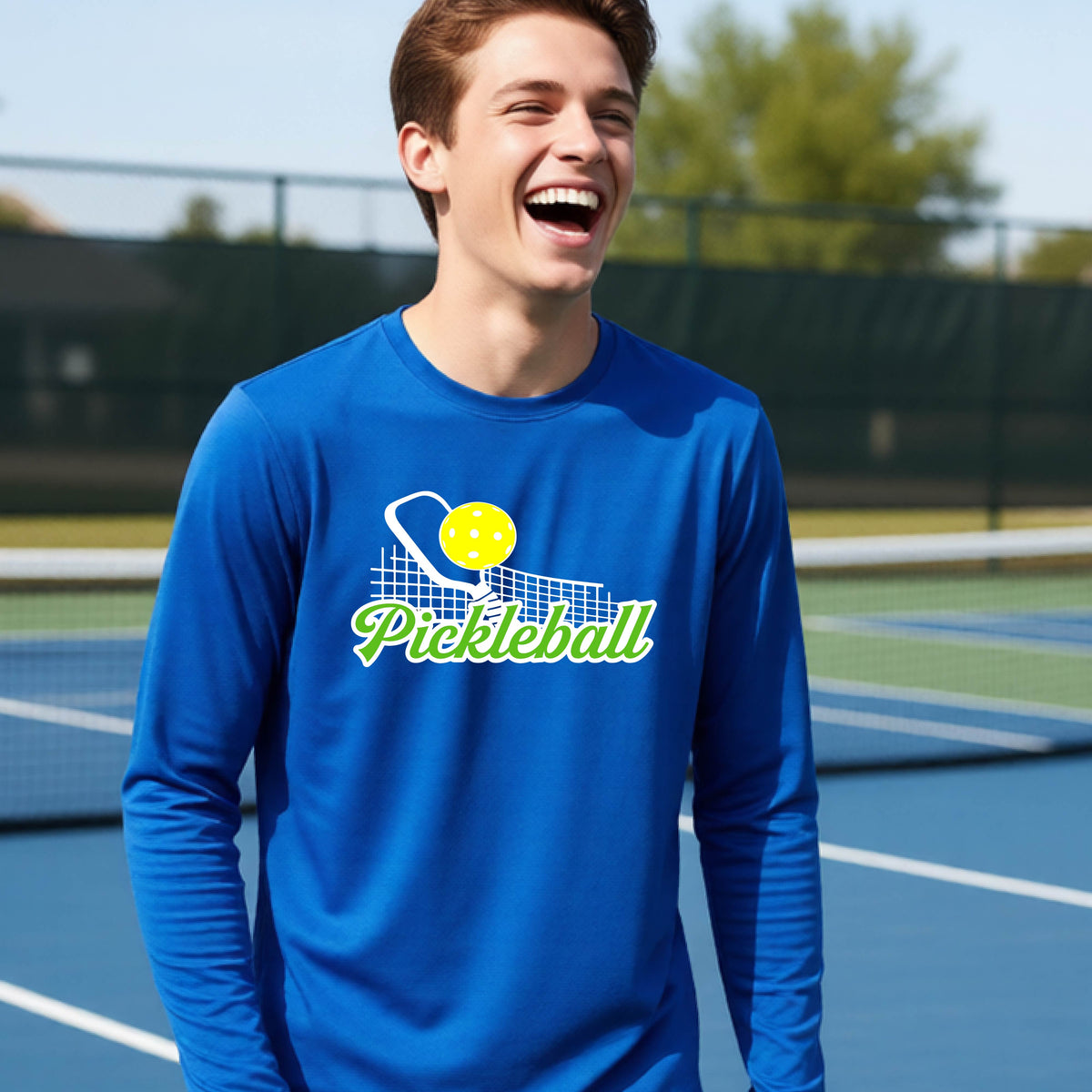 A young person wearing a royal blue long sleeve t-shirt with a graphic that has white and green text "PICKLEBALL'  with a white pickleball paddle and net and a yellow pickleball above the text.