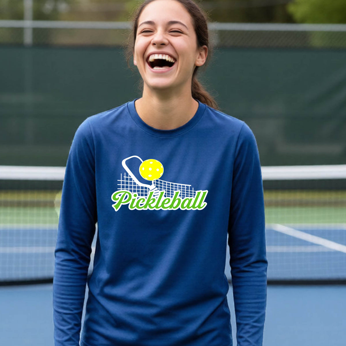  A young person wearing a royal blue long sleeve t-shirt with a graphic that has white and green text "PICKLEBALL'  with a white pickleball paddle and net and a yellow pickleball above the text.