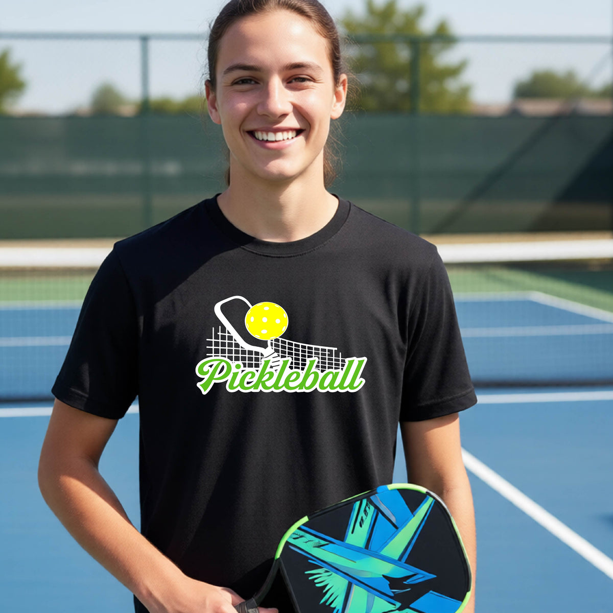A young person laughing on an outdoor pickleball court wearing a black t-shirt with a yellow and white pickleball graphic and the word 'Pickleball' printed on the front.
