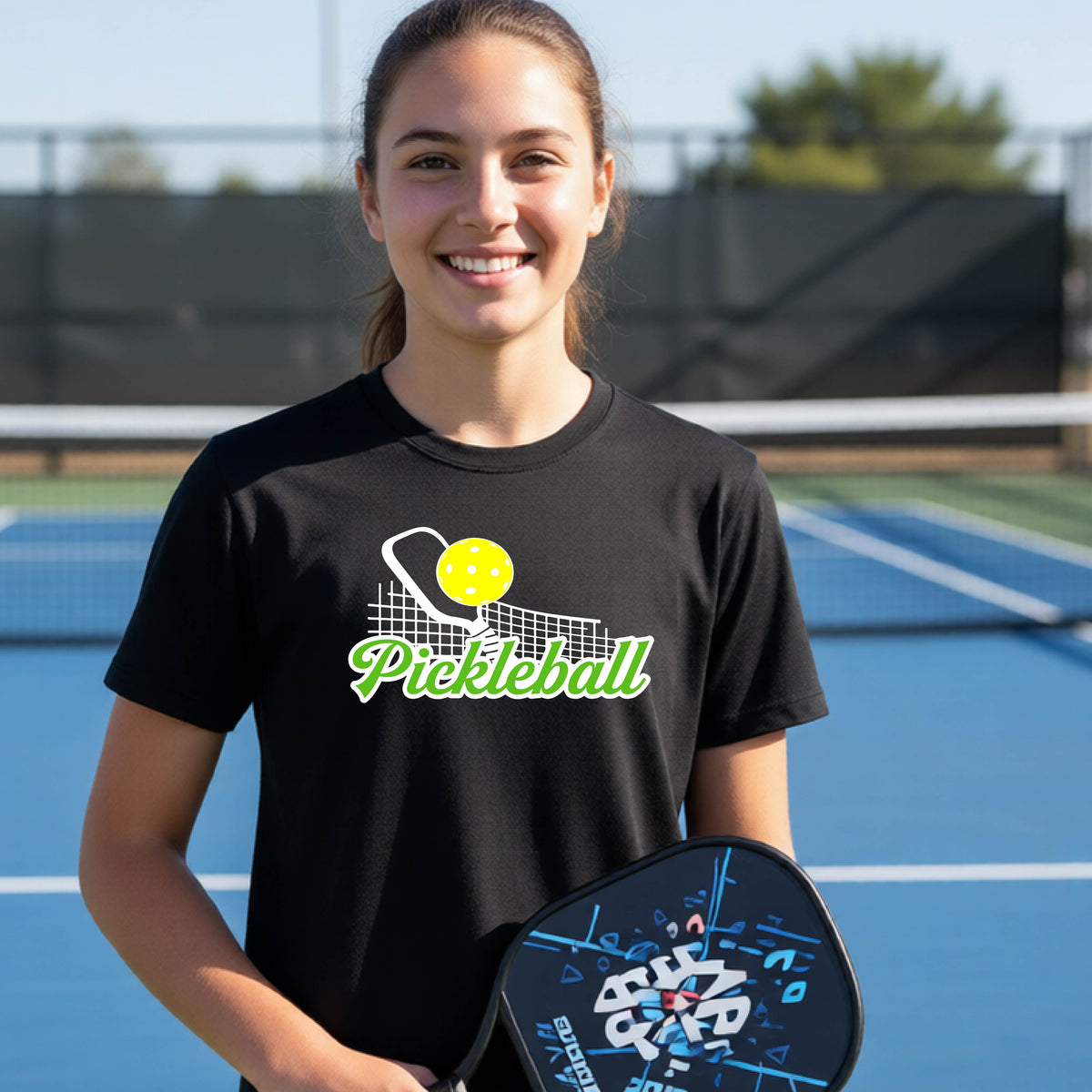 A young person laughing on an outdoor pickleball court wearing a black t-shirt with a yellow and white pickleball graphic and the word 'Pickleball' printed on the front.